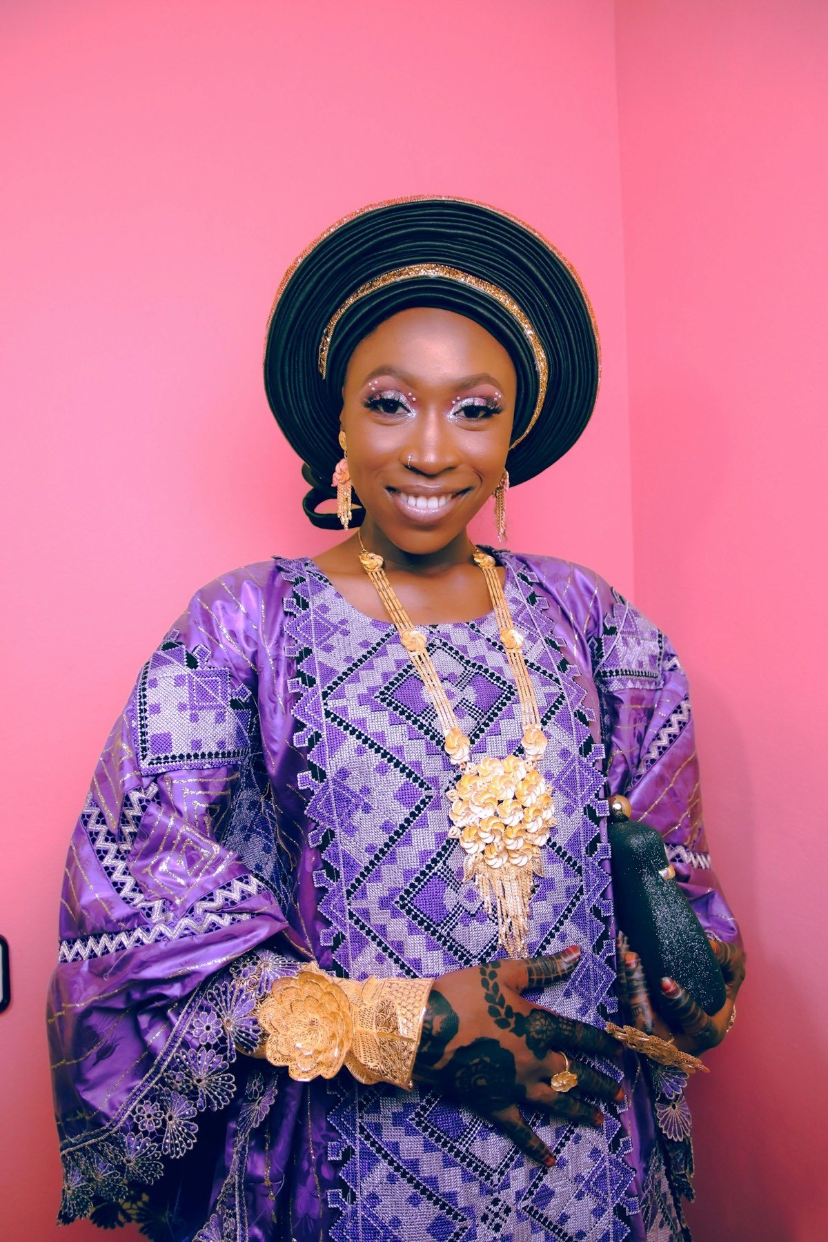 Designer-model in a purple patterned ensemble and wide-brimmed hat, standing against a pink wall.