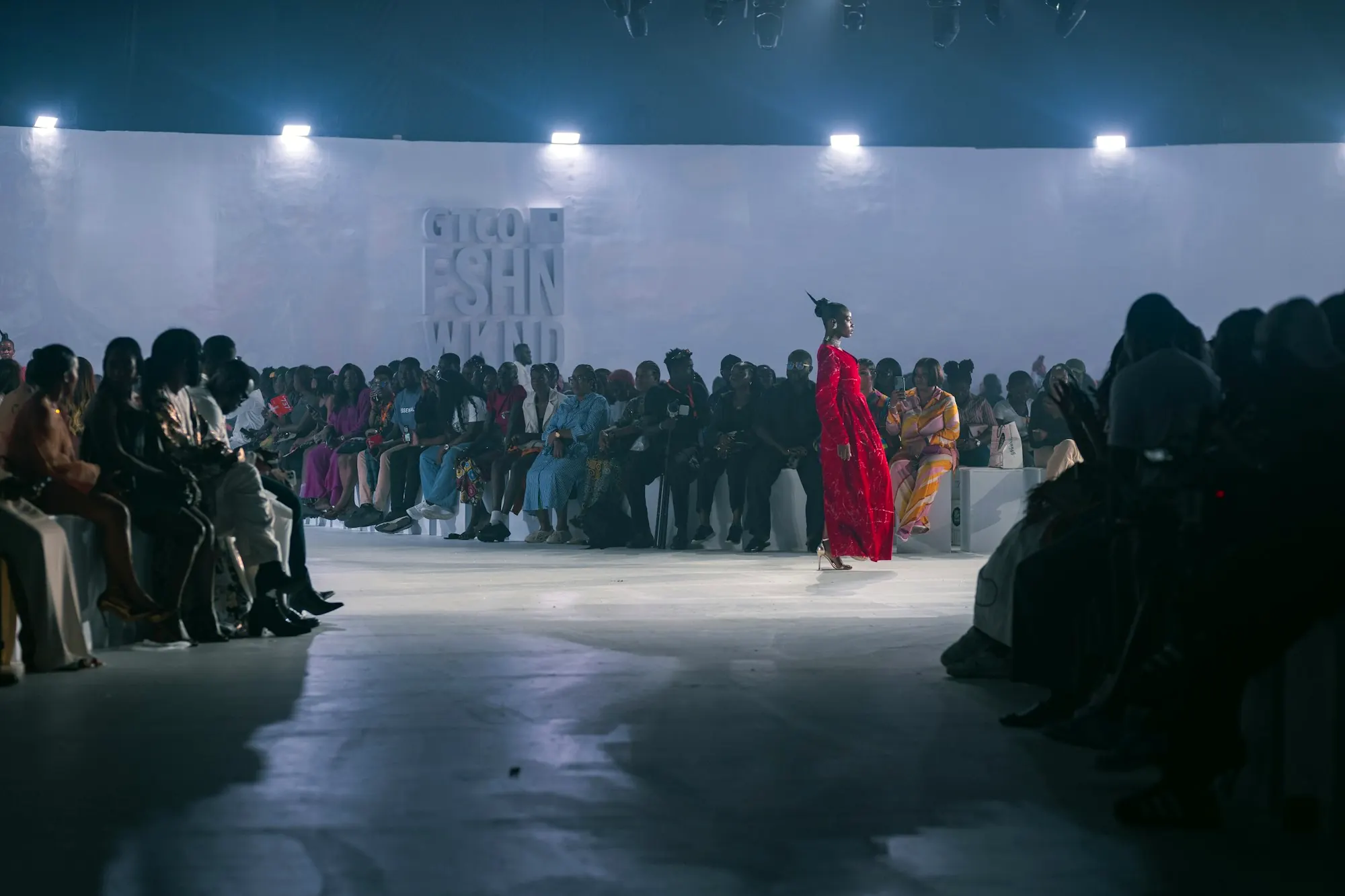 An audience seated along a white runway as models walk in the distance.