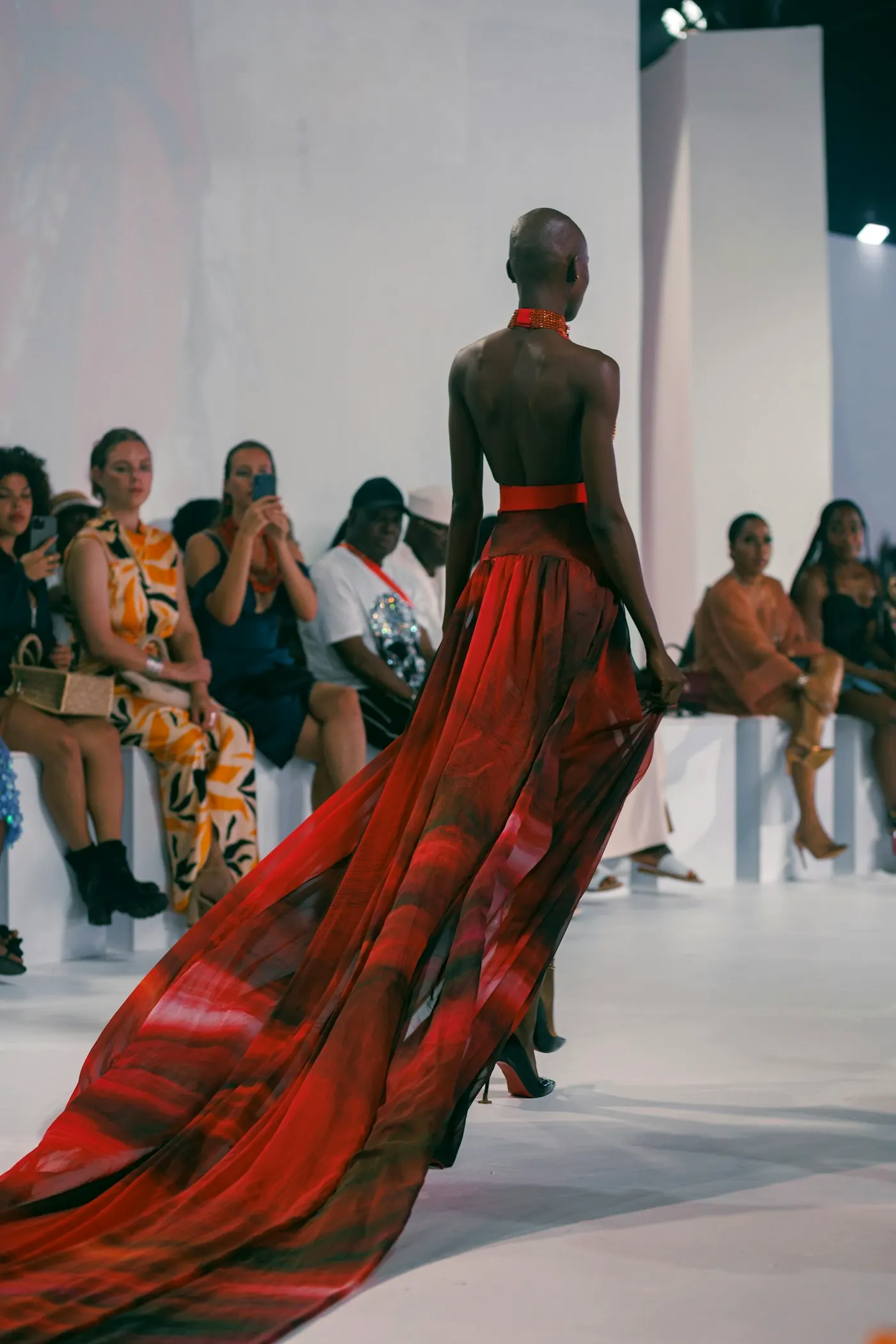 Model in a long red gown with a train, walking a white runway.